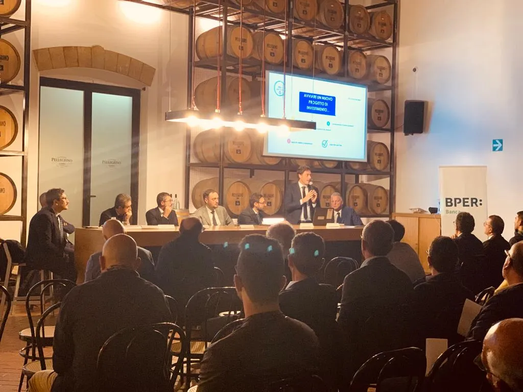 Nicola Parrinello in piedi al tavolo dei relatori, con il microfono, davanti alla sala; alle sue spalle una slide sull’avvio di un nuovo progetto di investimento e il pannello di BPER Banca