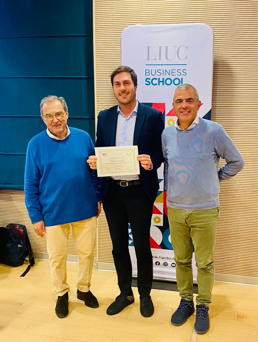 Nicola Parrinello con l’attestato in mano, tra due docenti, davanti al pannello della LIUC Business School