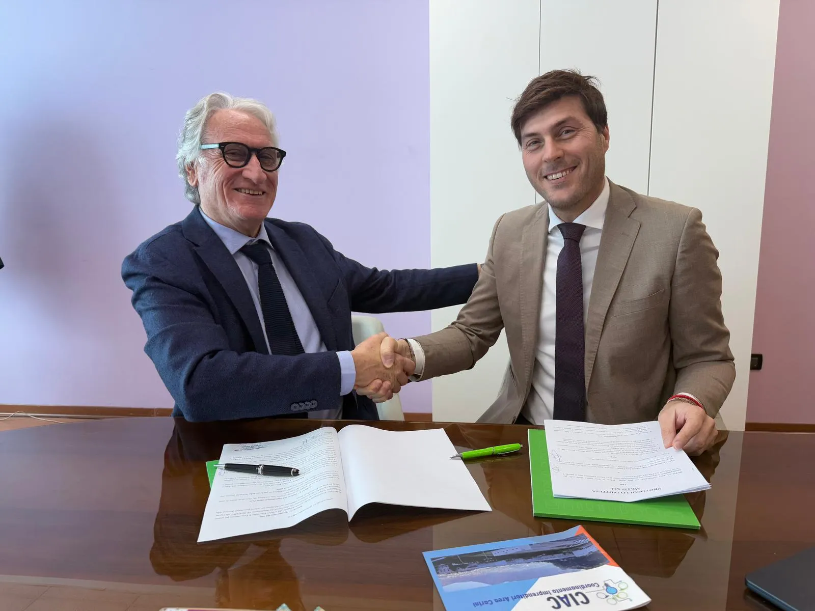 Nicola Parrinello e il rappresentante dell’associazione si stringono la mano guardando verso l’obiettivo; Nicola Parrinello tiene in mano il protocollo d’intesa